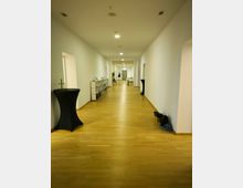 Langer, heller Flur mit hellem Holzfußboden und weißen Wänden. Entlang der Wände befinden sich ein hohes Bücherregal, Stehtische und vereinzelt Gegenstände wie Jacken oder Taschen; am Ende des Flurs ist eine geschlossene Tür sichtbar.