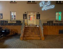 Straßenansicht eines Gebäudes mit der Leuchtschrift „Road House“ an der Wand. Eine Treppe aus roten Ziegelsteinen führt zu einer weißen Eingangstür, flankiert von Metallgeländern; daneben befinden sich Schilder mit der Aufschrift „Eventhouse“.