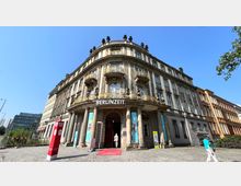 Historisches Gebäude mit mehreren Etagen, reich verzierten Balkonen und Säulen am Eingang, beschriftet mit "BERLINZEIT". Vor dem Eingang ist ein roter Teppich, und im Vordergrund befinden sich ein Besucher und Infotafeln.