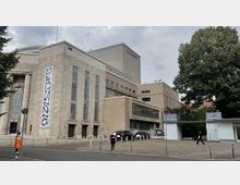 Historisches Gebäude mit heller Steinfassade und hohen rechteckigen Fenstern, ergänzt durch modernere Anbauten mit flacheren Linien und Glasflächen. Im Vordergrund ein kleiner Parkplatz, Bäume und Gehwege; grauer Himmel im Hintergrund.