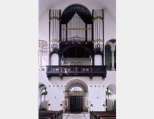 Innenraum einer Kirche mit Blick auf eine große Orgel, die sich über einer dunklen Holzempore befindet. Der Raum ist von weißen Wänden, hohen Bögen und symmetrischen Architekturmerkmalen geprägt.