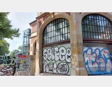Rote Backsteinfassade mit großen, bogenförmigen Fenstern und bemalten Rolltoren, bedeckt von Graffitis. Im Hintergrund befindet sich eine moderne verglaste Aufzugsanlage, umgeben von Bäumen.