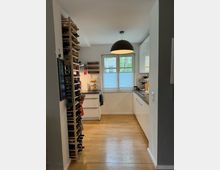 Moderne Küche mit Holzfußboden, einem großen Fenster mit Sichtschutz, weißen Einbauschränken und Arbeitsflächen. Links steht ein Regal mit Weinflaschen, und eine schwarze Hängelampe hängt zentral von der Decke.