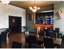 Ein modernes Restaurantinnere mit dunklen Holztischen und gepolsterten Stühlen. Im Hintergrund befindet sich eine Bar mit beleuchteten Getränkeregalen, daneben eine Tür in einen angrenzenden Raum und eine schlichte Wanddekoration.