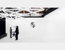 Ein minimalistisches Fotostudio mit weißen Wänden und Boden, einer Deckeninstallation aus kreuzförmigen, leuchtenden LED-Röhren. Zwei Personen stehen im Raum: eine Person hält eine Kameraausrüstung und filmt eine andere Person. Ein schwebendes metallisches Smiley-Objekt ist in der Mitte des Raumes sichtbar.