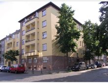 Wohngebäude in einer städtischen Umgebung, mit hellgelber Fassade und rotbraunem Sockelbereich aus Backstein; im Vordergrund parken Autos entlang einer gepflasterten Straße, und Bäume säumen den Gehweg.