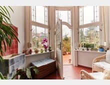 Heller Innenbereich mit großen Fensterflächen und dekorativem Fensterglas, geöffnetem Türbereich zum Außenbereich mit roten Terrakotta-Fliesen, Korbmöbeln und einer Sitzbank. Pflanzen und dekorative Objekte schmücken die Fensterbank, eine Wand ist mit roten Kacheln verkleidet.