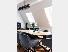 Esszimmer mit modernem Design: Ein rechteckiger Holztisch mit schwarzen Tischbeinen, umgeben von grauen, gepolsterten Stühlen. Darüber hängt eine Lampe mit mehreren schwarzen Glasleuchten, und zwei Dachfenster sorgen für natürliches Licht.