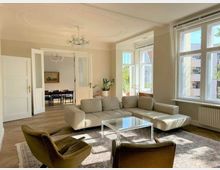 Helles Wohnzimmer mit großen Fenstern, einem beigen Ecksofa, zwei Sesseln und einem Glastisch auf einem Teppich. Im Hintergrund ist ein angrenzender Essbereich mit einem Tisch und Stühlen durch geöffnete Doppeltüren sichtbar.