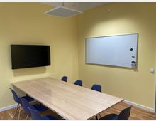 Konferenzraum mit gelb gestrichenen Wänden, einem rechteckigen Holztisch, umgeben von blauen Stühlen. An der Wand hängen ein Flachbildschirm und ein Whiteboard mit Markern in einer Halterung.