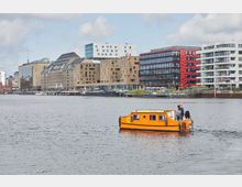 Eine städtische Szenerie mit modernen Gebäuden und sanierten Backsteinbauten entlang eines Flusses, im Vordergrund ein kleines, leuchtend orangefarbenes Boot mit zwei Personen an Bord.