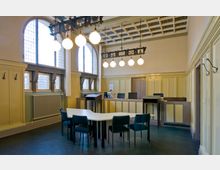 Historischer Sitzungssaal mit hohen, vergitterten Fenstern, gelb gestrichenen Holzvertäfelungen und einer dekorativen Deckenbeleuchtung aus kugelförmigen Leuchten. Im Vordergrund stehen Tische und blaue Stühle, im Hintergrund befindet sich eine erhöhte Richterbank.