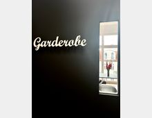 Schwarze Wand mit dem Schriftzug „Garderobe“ in weißer Schrift. Rechts ein schmales Fenster, durch das ein Innenraum mit einem Waschbecken und eine Vase mit roten Blumen sowie ein Gebäude mit weißer Fassade sichtbar sind.