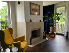 Helles Wohnzimmer mit dunklem Holzboden und einem offenen, hell verkleideten Kamin in der Raumecke. Große Fenster mit Vorhängen geben den Blick ins Grüne frei; davor stehen ein gelber Sessel, eine mehrkugelige Stehlampe und eine große Zimmerpflanze auf einem kleinen Tisch.
