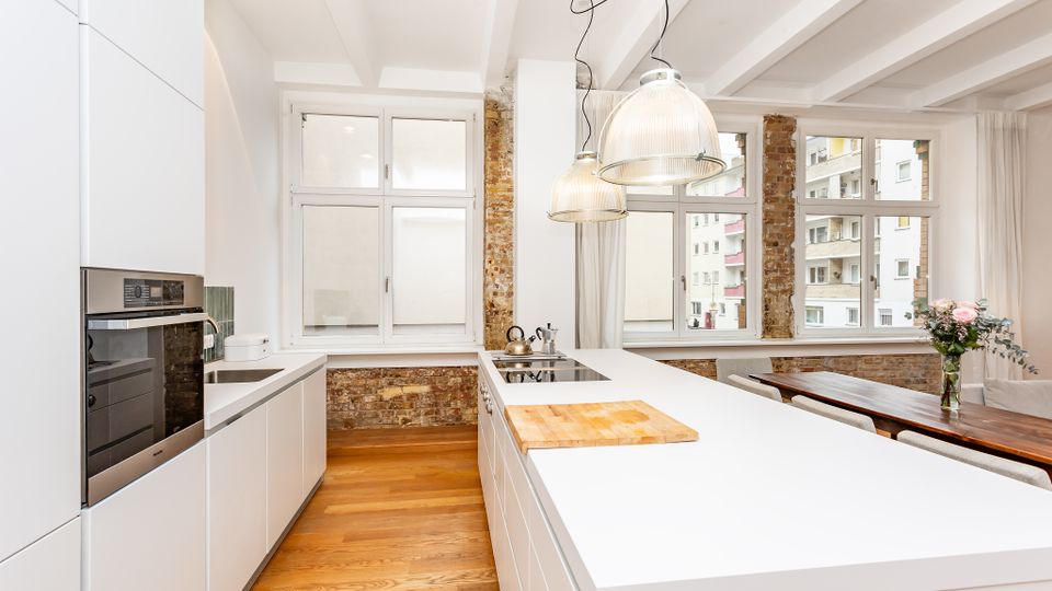 Helle, moderne Wohnküche mit weißen Einbauten und großer Kücheninsel, Holzfußboden und sichtbaren Backsteinpfeilern. Große Fensterfronten und zwei hängende Glaslampen beleuchten den Raum; daneben steht ein Esstisch am Fenster.