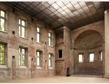 Innenraum eines historischen Backsteingebäudes mit hohen Wänden, großen Fenstern und sichtbarer Bausubstanz. Eine abgerundete Apsis ist auf der rechten Seite sichtbar, und die Decke besteht aus einem modernen, transparenten Material, das Licht hereinlässt.