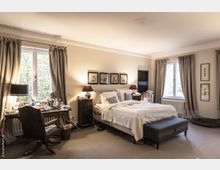 Helles Schlafzimmer mit einem großen Doppelbett, flankiert von Nachttischen mit Lampen. Links ein Schreibtisch mit Stuhl vor einem Fenster mit Vorhängen, rechts weitere Fenster mit Blick nach draußen; Bücherstapel und Wandbilder als Dekoration.