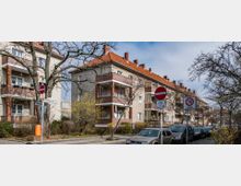 Straßenszene in einer Wohngegend mit dreigeschossigen Mehrfamilienhäusern aus Backstein und Putz, roten Ziegeldächern und kleinen Balkonen. Im Vordergrund sind Straßenschilder, parkende Autos und kahle Bäume zu sehen, die eine Verkehrszone mit 30 km/h markieren.