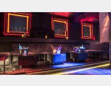 Ein stilvoller Innenraum einer Lounge oder Bar mit dunklen Sitzbänken, niedrigen Tischen und beleuchteten Flaschen in Eiskübeln. Neonbeleuchtete quadratische Rahmen an den Wänden verleihen dem Raum eine moderne Atmosphäre.