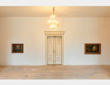 Heller Galerieraum mit weißer Wand, mittig einer hohen, klassisch gerahmten Doppeltür und darüber einem Kronleuchter. Links und rechts hängen gerahmte Landschaftsgemälde, der Boden ist roh und unbedeckt.