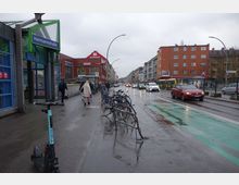 Straßenszene in einer urbanen Umgebung mit dem Eingang zur U-Bahn-Station "Hermannstraße" auf der linken Seite, mehreren geparkten Fahrrädern, einem Roller im Vordergrund und Autos sowie Bussen auf einer nassen, regnerischen Fahrbahn. Im Hintergrund sind Geschäfte, darunter ein REWE-Supermarkt, und mehrstöckige Wohnhäuser zu sehen.