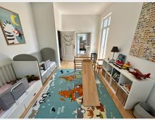 Helles Kinderzimmer mit zwei Betten an der linken Seite, einem bunten Teppich mit Tiermotiven auf dem Boden und einer langen Holzrampe in der Mitte. Rechts befinden sich Regale mit Spielsachen und Büchern, sowie ein kleiner Spieltisch vor einer großen Glastür.