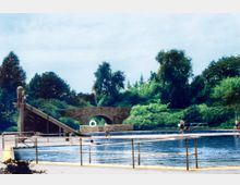 Freibad mit einem großen Schwimmbecken, einer Wasserrutsche und Geländern im Vordergrund; im Hintergrund ein steinerner Brückenbogen und dicht bewachsene grüne Bäume.