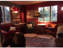 Gemütlicher Wohnraum mit roten Wänden, großen Fenstern und einer Mischung aus Vintage-Möbeln, darunter ein rotes Sofa, ein grüner und ein orangefarbener Sessel. Ein Teppich mit orientalischem Muster bedeckt den Boden, und der Raum wird von einer Stehlampe sowie Tageslicht durch die Fenster beleuchtet, mit Blick auf einen Garten.