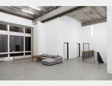 Offener, minimalistischer Innenraum mit weißen Wänden, einer grauen Couch und einem Holztisch auf grauem Boden. Links befindet sich eine Fensterfront mit hohen Fenstern, rechts führen Treppen mit schwarzem Geländer nach unten.