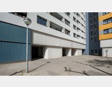 Ein moderner Wohnkomplex mit weißer, blauer und gelber Fassadengestaltung; im Vordergrund ein breiter Gehweg mit Betonplatten und eine Laterne. Unter dem Gebäude befinden sich offene Eingänge und Durchgänge, die von Betonwänden gestützt werden.