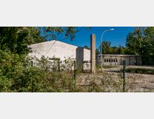 Industriegelände mit mehreren weiß gestrichenen, flachen Gebäuden aus Beton, umgeben von einer verrosteten Gitterzaunanlage und dichter Vegetation. Im Hintergrund stehen Bäume unter einem klaren blauen Himmel.