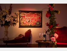 Ein geschmackvoll gestalteter Innenraum mit einem leuchtend roten Plakat an der Wand mit der Aufschrift "Tangoloft Berlin" in verschnörkelter Schrift. Im Vordergrund ein rotes Sofa mit goldenen Verzierungen, Blumenarrangements und ein Tisch mit Champagnerflasche und Gläsern.