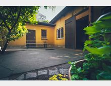 Ein kleiner Innenhof mit gelber Hausfassade, zwei Fenstern mit Metallgittern, einer geschlossenen Tür und einem offenen Scheunentor. Im Vordergrund stehen Pflanzen in einem Blumentopf, rechts ist eine Sitzbank erkennbar.