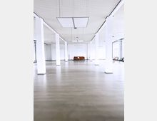 Großer, heller Raum mit weißen Wänden und vielen rechteckigen, weißen Säulen, die den Raum gliedern. Der Boden ist grau und glatt, die Decke perforiert mit rechteckigen Hängelampen; im Hintergrund steht ein einzelnes braunes Sofa.