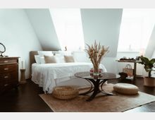 Helles Schlafzimmer mit schrägen Wänden und Dachfenstern, einem Bett mit weißer Bettwäsche, einem dunklen Holztisch mit getrockneten Gräsern in der Mitte, geflochtenen Sitzkissen auf einem Teppich sowie einem kleinen Holztisch mit Dekoration und einer Zimmerpflanze.