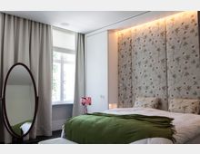 Schlafzimmer mit einem Bett, das mit weißen Kissen und einer grünen Decke ausgestattet ist, vor einer Wand mit floraler Tapete. Links steht ein ovaler Standspiegel, neben einem Fenster mit beigefarbenen Vorhängen und Lamellenrollo.