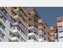 Mehrstöckiges Wohngebäude mit farbigen Fassaden in Braun-, Lila- und Weißtönen, das über zahlreiche Balkone verfügt. Die Fassade ist symmetrisch angeordnet, und Satellitenschüsseln sind an einigen der Balkone montiert; klarer blauer Himmel im Hintergrund.
