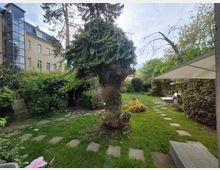Grüner Garten mit üppigen Sträuchern, einem großen Baum und gepflasterten Gehwegen. Im Hintergrund ist ein mehrstöckiges Wohngebäude mit modernen Glaselementen zu sehen; ein Sonnensegel und Gartenmöbel befinden sich rechts im Bild.