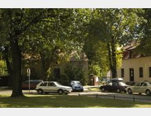 Ein kleiner, von Bäumen umgebener Platz mit parkenden Autos vor einem alten, verwachsenen Gebäude mit Ziegeldach. Rechts daneben steht ein modernes, helles Gebäude mit Fenstern und roten Dachelementen.