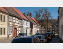 Straße mit parkenden Autos und historischen Fachwerkhäusern auf beiden Seiten, mit roten Ziegeldächern und weißen Fensterrahmen. Im Hintergrund sind ein weiteres Fachwerkhaus und kahle Bäume unter einem klaren blauen Himmel sichtbar.