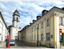 Straßenszene mit barockem Kirchturm im Hintergrund und hellgelben Gebäuden mit verzierten Fassaden im Vordergrund; rechts ein Schild mit einer Einfahrt-Verboten-Markierung, links ein Werbeaufsteller für das Museum Fürstenwalde.