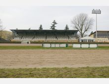 Eine Tribüne auf einem Pferderennplatz mit überdachten Sitzplätzen, flankiert von Bäumen und einem Beleuchtungsmast. Im Vordergrund sind eine Sandbahn, grüne Absperrungen und eine Rasenfläche sichtbar.