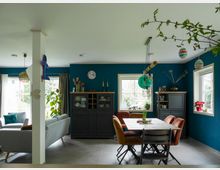 Wohnbereich in einem modernen Haus mit blauen Wänden und großen Fenstern, die viel Tageslicht einlassen. Der Raum ist mit einer Sitzecke, einem Esstisch mit farbigen Stühlen und einer dunklen Anrichte eingerichtet, dekoriert mit Zimmerpflanzen und hängenden Ornamenten.