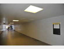 Ein beleuchteter, gefliester Fußgängertunnel mit weiß gekachelten Wänden, einem gelblichen Informationsaushang rechts und zwei Personen, die am Ende des Tunnels in Richtung Tageslicht gehen.