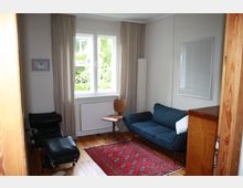 Kleines Wohnzimmer mit Holzdielenboden, einem roten Teppich, einem blauen Sofa, einem Holzdrehstuhl und einem schwarzen Liegesessel. Ein Fenster mit beigen Vorhängen und Blick ins Grüne erhellt den Raum, an der Wand hängt ein kleines Bild.