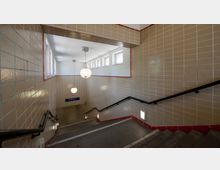 Eine Treppe in einem U-Bahnhof mit beigen Wandfliesen, roten Fliesenabschlüssen und schwarzen Handläufen. Oben befinden sich kleine Fenster und hängende Kugelleuchten, die den Bereich beleuchten.