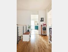 Flur eines modernen Wohnhauses mit hellem Holzboden, einer weißen Sitzbank mit integrierten Aufbewahrungsfächern links, und Regalmöbeln rechts. Im Hintergrund führt eine offene Tür in ein helles Zimmer mit grüner Kommode und großen Fenstern.