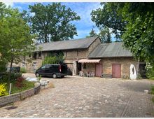 Ein gepflasterter Hof mit einem historischen Backsteingebäude, das teils mit Efeu bewachsen ist. Rechts sind rote Holztüren und unter einem gestreiften Sonnendach stehen Gartentische mit Stühlen; im Vordergrund ein parkender schwarzer Van und grünes Gebüsch um den Hof.