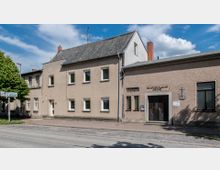 Straßenszene mit einem schlichten, zweigeschossigen Wohngebäude aus Putzfassade und einem flacheren angrenzenden Gebäude mit der Aufschrift „Neuapostolische Kirche“ über dem Eingang. Im Vordergrund ein Gehweg und eine Straße mit einem Wegweiser, daneben steht ein Baum.