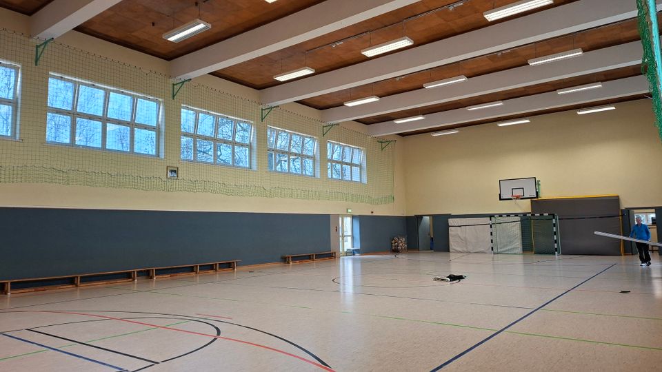 Innenansicht einer großen Sporthalle mit markiertem Hallenboden, hohen Fensterreihen entlang der Seitenwand und einer Holzdecke mit langen Deckenleuchten. An der Stirnseite sind ein Basketballkorb und ein Handballtor vor gepolsterten Wandflächen sowie Sitzbänke entlang der Wand zu sehen.